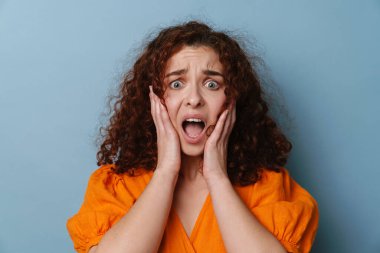 Şok olmuş kıvırcık kız mavi arka planda mahsur kalmış kamerada şaşkınlığını dile getiriyor.