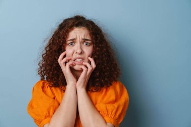 Yüzünü buruşturan ve yüzüne dokunan ürkek kızıl kadın mavi arka planda izole edilmiş.