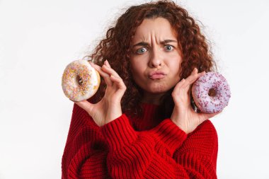 Kızıl kıvırcık kız beyaz arka planda izole edilmiş çöreklerle dalga geçerken yüzünü buruşturuyor.