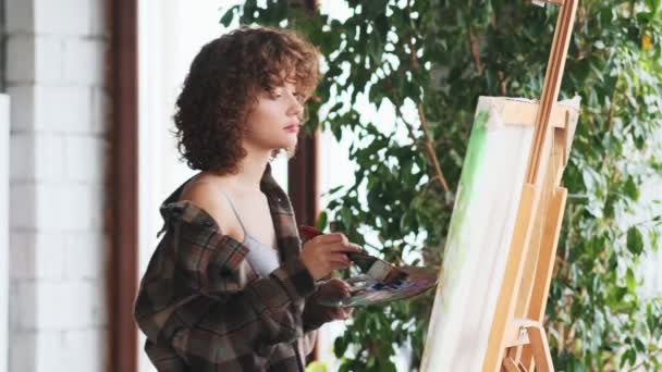 一个迷人的女艺术家站在画架旁边 在她的画室里画着一幅油画 — 图库视频影像