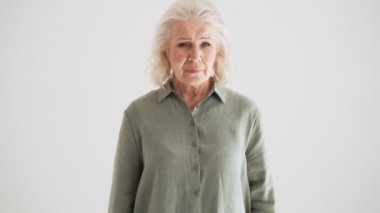 Hayal kırıklığına uğramış yaşlı bir kadın, elini sallarken kameraya poz veriyor. Stüdyonun beyaz duvarında izole bir şekilde duruyor.