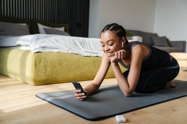 Attractive young smiling african woman listening to music with earphones in the bedroom, using mobile phone