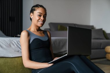 Healthy young fit african sportswoman using laptop computer while sitting on a fitness mat in the bedroom