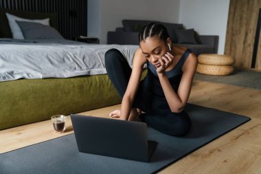 Healthy young fit african sportswoman using laptop computer while sitting on a fitness mat in the bedroom