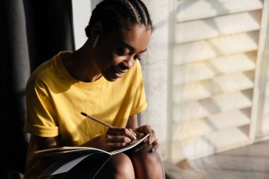 Happy african american girl in earphones writing down notes while sitting at window in home