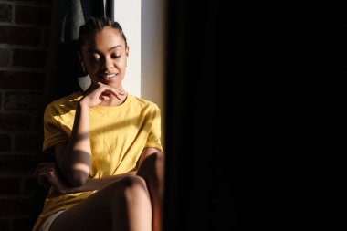 Pleased african american girl listening music with earphones while sitting at window in home