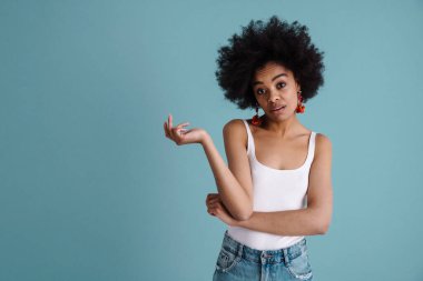 Kafası karışmış Afrikalı Amerikalı kız mavi arka planda yalnız poz veriyor ve kameraya bakıyor.