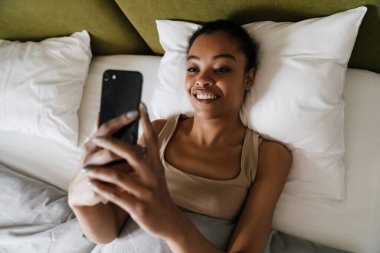 Mutlu Afro-Amerikan kızı evde uyuduktan sonra yatakta cep telefonu kullanıyor.