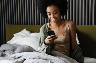 Mutlu Afro-Amerikan kızı evde uyuduktan sonra yatakta cep telefonu kullanıyor.