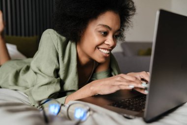 Orta yaşlı Afrikalı kadın evde yatarken dizüstü bilgisayar kullanıyor.