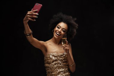 Mutlu Afrikalı genç bir kadın stüdyoda cep telefonuyla selfie çekiyor, elinde bir kadeh şampanya tutuyor.