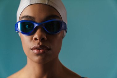 Mavi duvarın üzerinde duran mayolu ve yüzme şapkalı Afrikalı genç bir kadının portresini yakından çek.