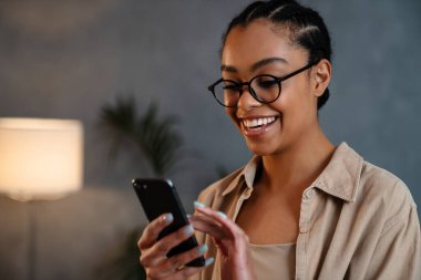 Neşeli Afrikalı Amerikalı kadın cep telefonu kullanıyor ve içeride gülümsüyor.