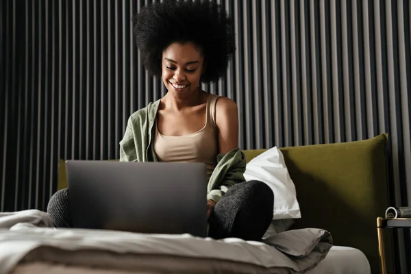 坐在床上用笔记本电脑笑着的非洲裔美国年轻女人 — 图库照片