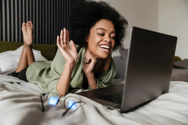 快乐的中年非洲女人在床上躺着的时候用笔记本电脑 在家里自我隔离的时候用视频聊天 — 图库照片