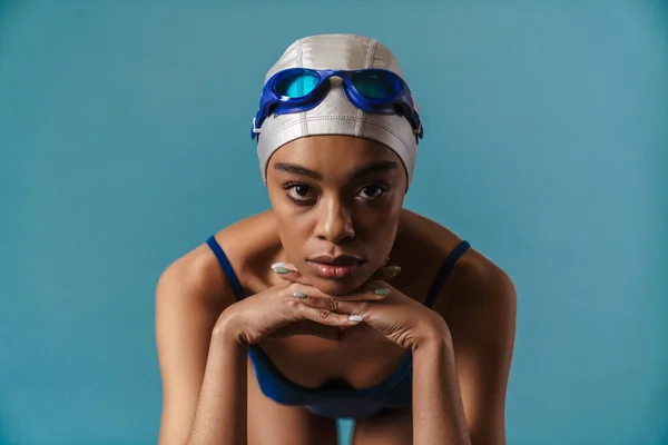Young black swimmer in goggles posing and looking at camera isolated over blue wall