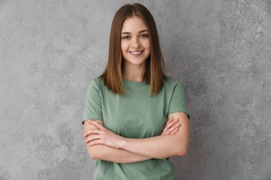 Mutlu güzel kız kollarını kavuşturup gri duvarın üzerinde yalnız poz veriyor.