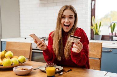 Mutfak masasında oturmuş internetten alışveriş yapmak için cep telefonu ve kredi kartı kullanan neşeli genç bir kadın.