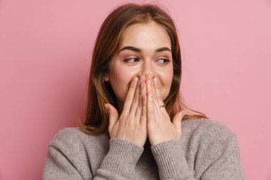 Genç kızıl mutlu kadın pembe arka planda ağzını kapatırken yana bakıyor.