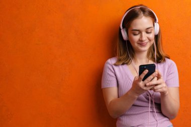 Turuncu duvarda izole edilmiş kulaklıkları ve cep telefonlarıyla müzik dinleyen kızıl saçlı genç bir kadın.