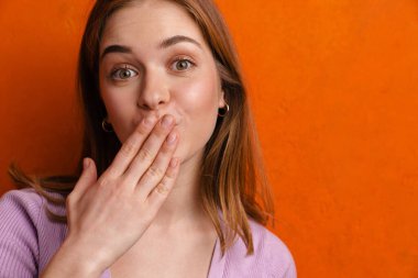 Genç kızıl mutlu kadın, turuncu duvarın üzerinden ağzını kapatırken kameraya bakıyor.