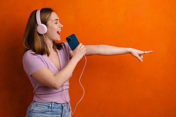 Joyful Woman Using Headphones Cellphone While Pointing Finger Camera Isolated — 스톡 사진