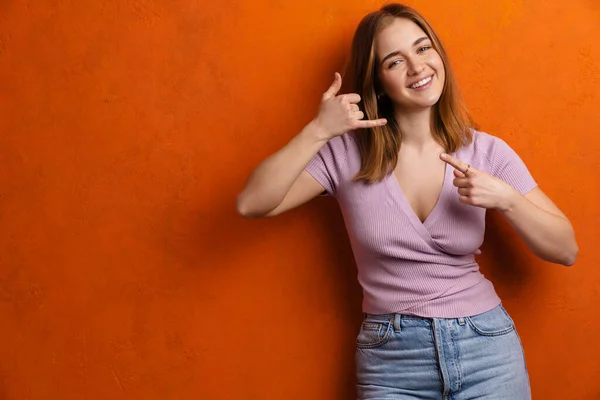 Uśmiechnięta Młoda Dziewczyna Pokazuje Telefon Gest Izolowany Pomarańczowym Tle Wskazując — Zdjęcie stockowe