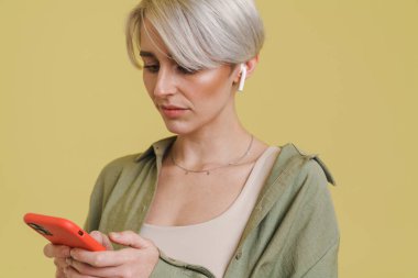 Bej arka planda izole edilmiş cep telefonu ve kulaklıklarıyla dinleyen genç gri kadın.