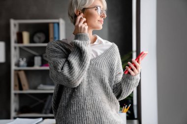Ofiste dururken cep telefonu ve kulaklık kullanan beyaz saçlı bir kadın.