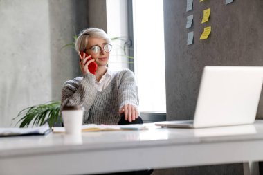 Ofiste dizüstü bilgisayarla çalışırken cep telefonuyla konuşan beyaz saçlı kadın.