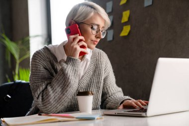 Ofiste dizüstü bilgisayarla çalışırken cep telefonuyla konuşan beyaz saçlı kadın.