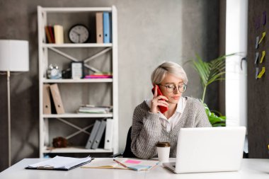 Ofiste dizüstü bilgisayarla çalışırken cep telefonuyla konuşan beyaz saçlı kadın.