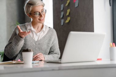 Beyaz saçlı, gülümseyen kadın, ofiste dizüstü bilgisayarla çalışırken el kol hareketi yapıyor.