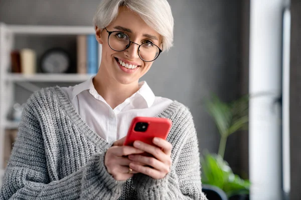 オフィスにいる間に携帯電話を使う白髪の笑顔の女性 — ストック写真