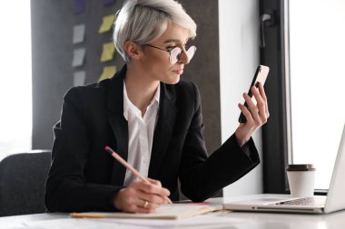 Ofiste dizüstü bilgisayarla çalışırken cep telefonu kullanan beyaz saçlı kadın.