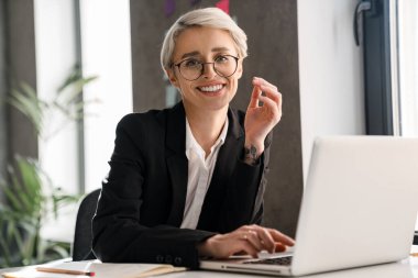 Ofiste otururken dizüstü bilgisayarla çalışan beyaz saçlı neşeli kadın.