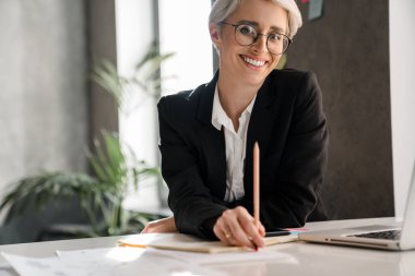 Ofiste otururken dizüstü bilgisayarla çalışan beyaz saçlı neşeli kadın.