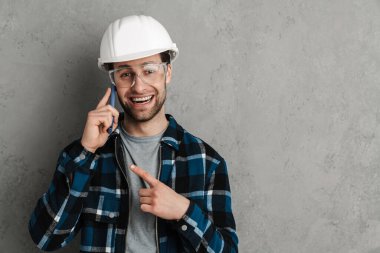 Neşeli müteahhit cep telefonuyla konuşuyor ve gri duvarda izole edilmiş bir şekilde işaret ediyor.