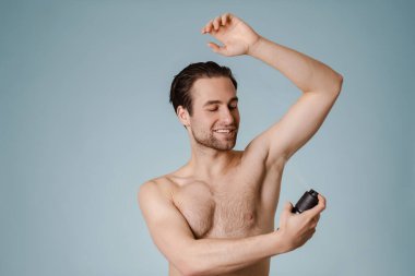 Yakışıklı, gülümseyen çıplak adam mavi arka planda izole edilmiş deodorant püskürtüyor.
