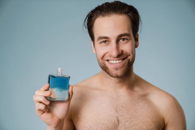 Üstsüz beyaz adam gülümserken mavi duvarın üzerinde izole edilmiş kolonya gösteriyor.