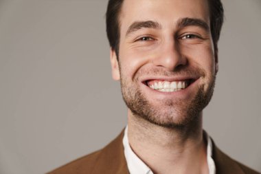 Tıraşsız esmer adam gülümsüyor ve gri arka planda izole edilmiş kameraya bakıyor.