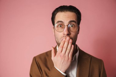 Şok olmuş esmer ceketli adam pembe duvarın arkasından kameraya bakıyor, ağzını kapatıyor.