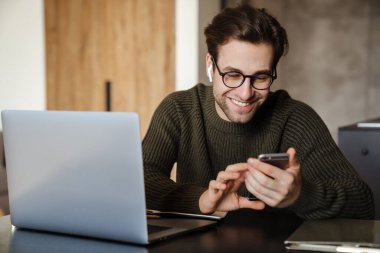 Gülümseyen esmer adam evde otururken telefon ve dizüstü bilgisayar kullanıyor.