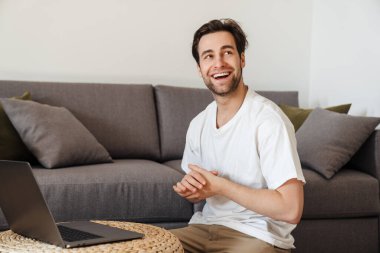 Gülümseyen esmer adam dizüstü bilgisayarla yerde oturuyor, başka tarafa bakıyor.