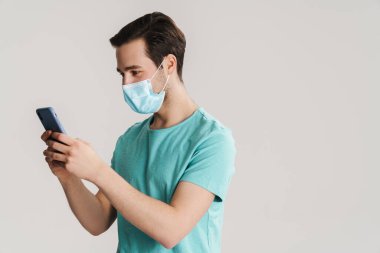Beyaz duvarda izole edilmiş cep telefonu kullanan koruyucu maskeli yakışıklı adam.