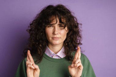 Kara kara düşünen esmer, kıvırcık kız parmaklarını çapraz olarak mor arka planda tutuyor.