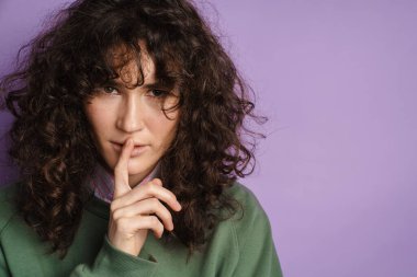 Genç, güzel kıvırcık kız sessiz hareketler yapıyor. Parmağı dudaklarında, mor arka planda.