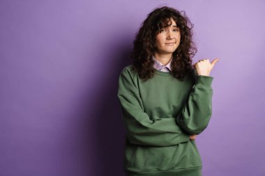 Genç, güzel kıvırcık kız, mor arka planda soyutlanmış bir şekilde parmağını yana gösteriyor.