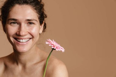 Bej arka planda pembe çiçekle poz verirken yarı çıplak kıvırcık kadın gülüyor.