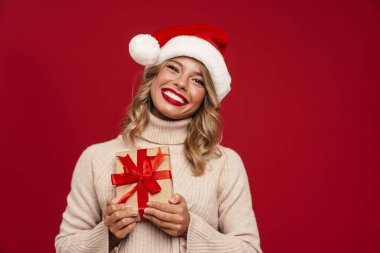 Kırmızı arka planda saklanmış hediye kutusunu tutan süveter giyen mutlu genç bir kadın.